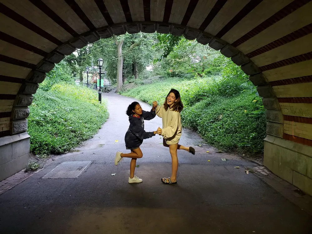 Two people are joyfully posing and smiling in a playful stance under a stone archway in a lush park setting
