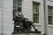 The image shows a bronze statue of a seated figure with a book on their lap, set against the backdrop of a building with large windows.
