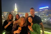 A group of people is posing with drinks on a rooftop at night with a brightly lit city skyline in the background.