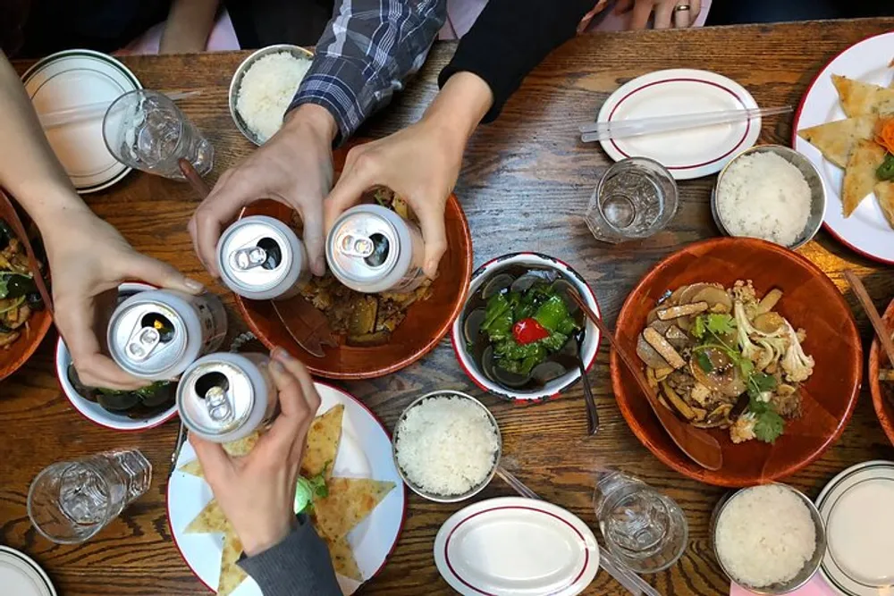 A group of people is toasting with canned drinks over a table filled with various dishes including rice and what appears to be Asian cuisine in a casual dining setting