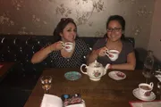 Two individuals are smiling and holding teacups in a cozy setting with vintage wallpaper and a teapot on the table, suggesting a casual and friendly tea time.