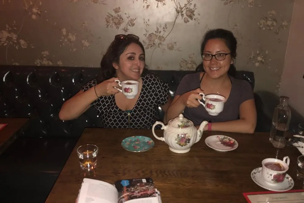 Two individuals are smiling and holding teacups in a cozy setting with vintage wallpaper and a teapot on the table suggesting a casual and friendly tea time
