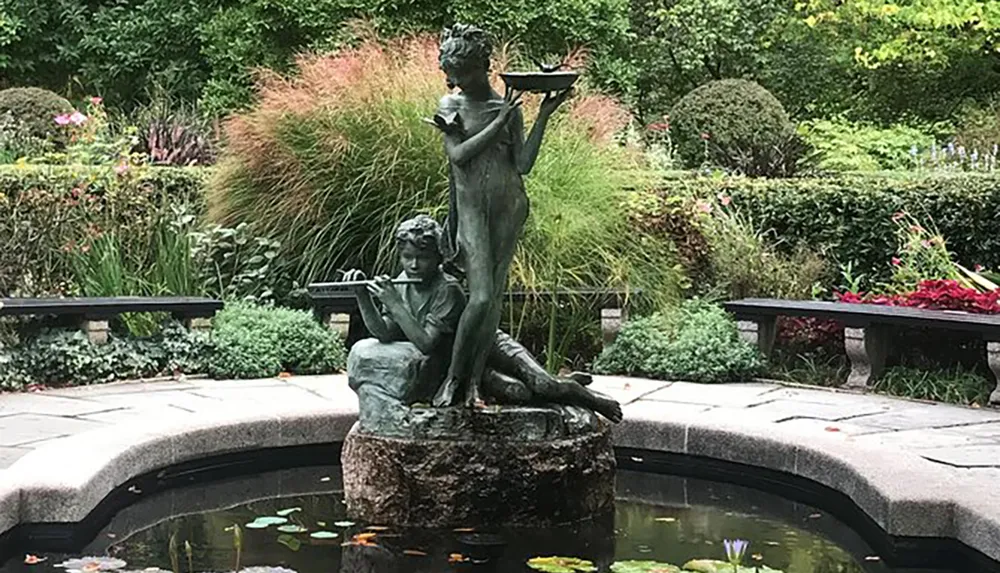 The image shows a serene garden featuring a bronze statue of two children one standing and holding a bird bath and the other seated and playing a flute set in the middle of a circular pond surrounded by lush plants and benches