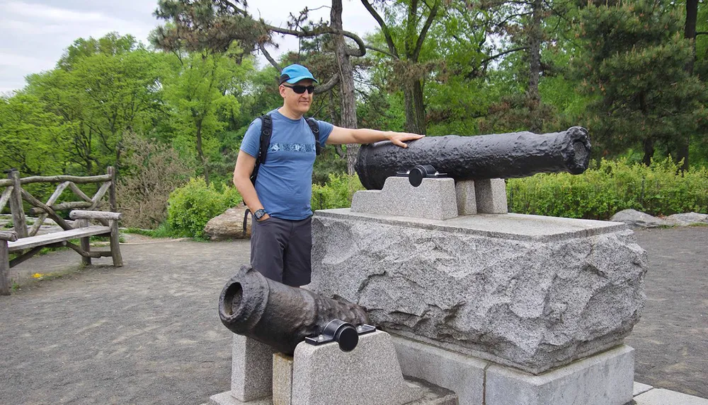 A man wearing a cap sunglasses and a backpack is smiling and standing next to an old cannon mounted on a stone base in an outdoor setting with trees in the background