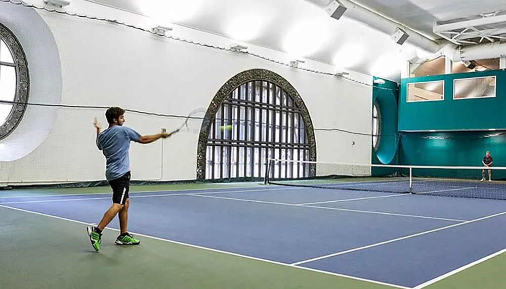 A person is playing tennis on an indoor court with a distinctive large round window and an arched gate on the wall behind