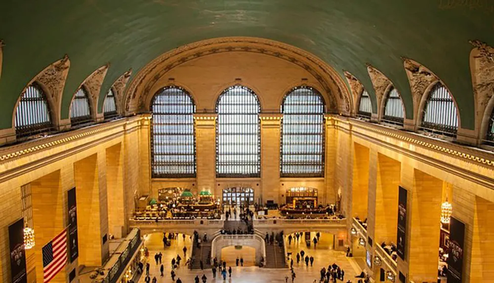 The image shows the spacious interior of a grand train station featuring large arched windows and a vaulted ceiling bustling with people and adorned with an American flag