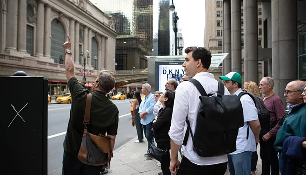 A group of people are standing on a city sidewalk with one man pointing upwards possibly on a tour or observing something of interest