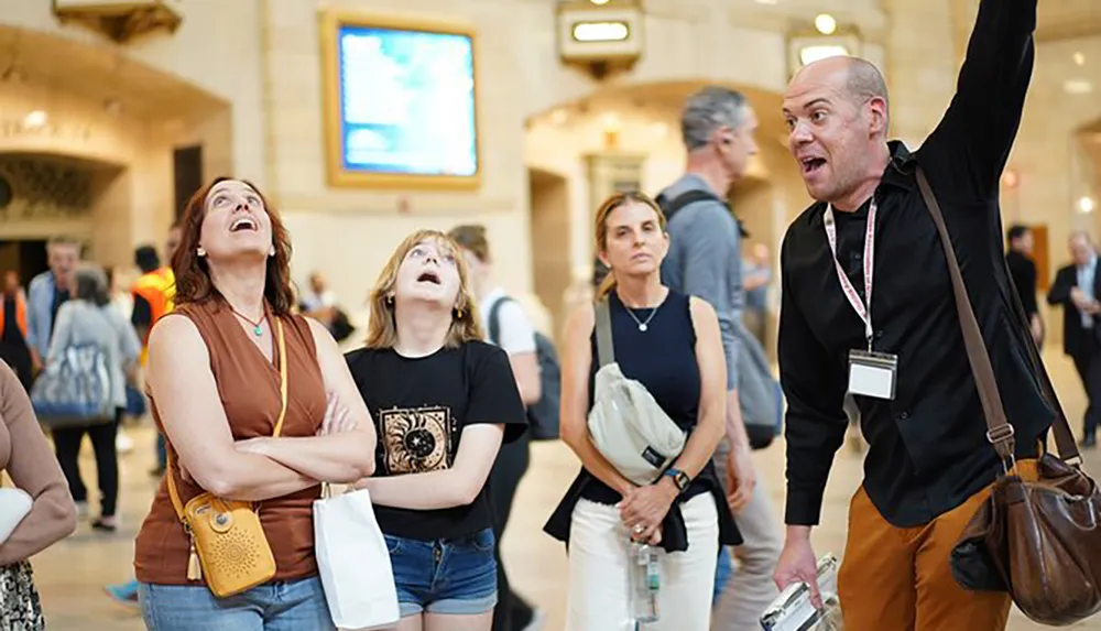 A group of tourists led by a gesticulating guide look upwards with interest inside a busy station or public space