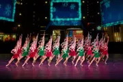 A group of dancers dressed in festive red and green costumes is performing a synchronized routine on a stage with a wintery, holiday-themed backdrop.