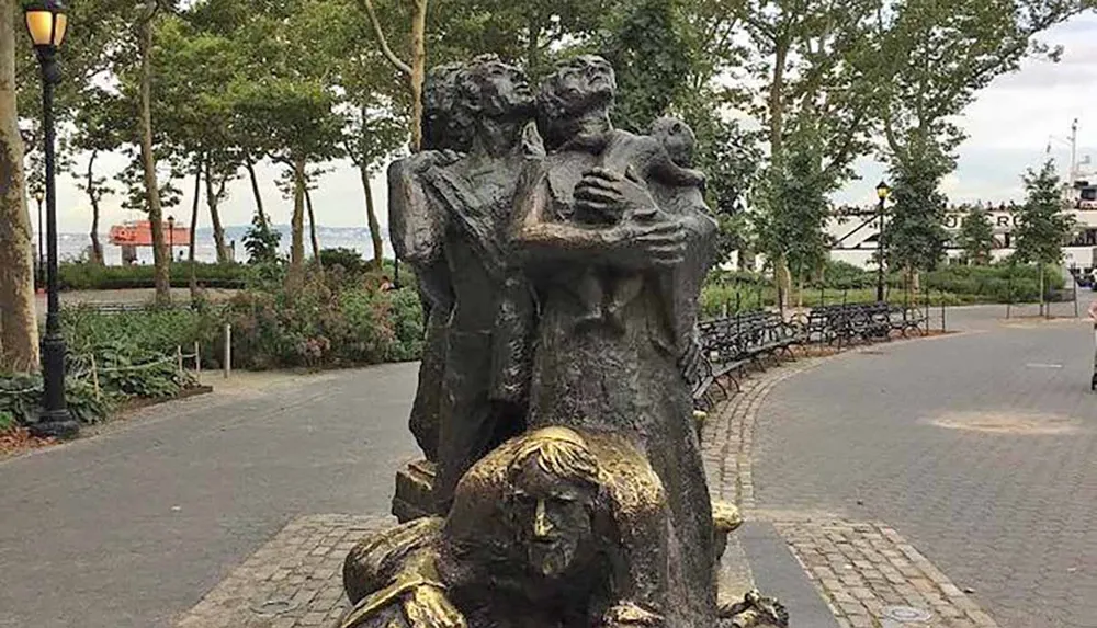 The image shows a bronze sculpture of a family with two adults and a child embraced in a close huddle set in a park with walkways and trees and a water body visible in the background