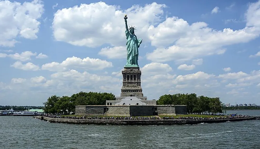 The image shows the Statue of Liberty against a backdrop of clouds and blue sky with visitors visible on the ground nearby