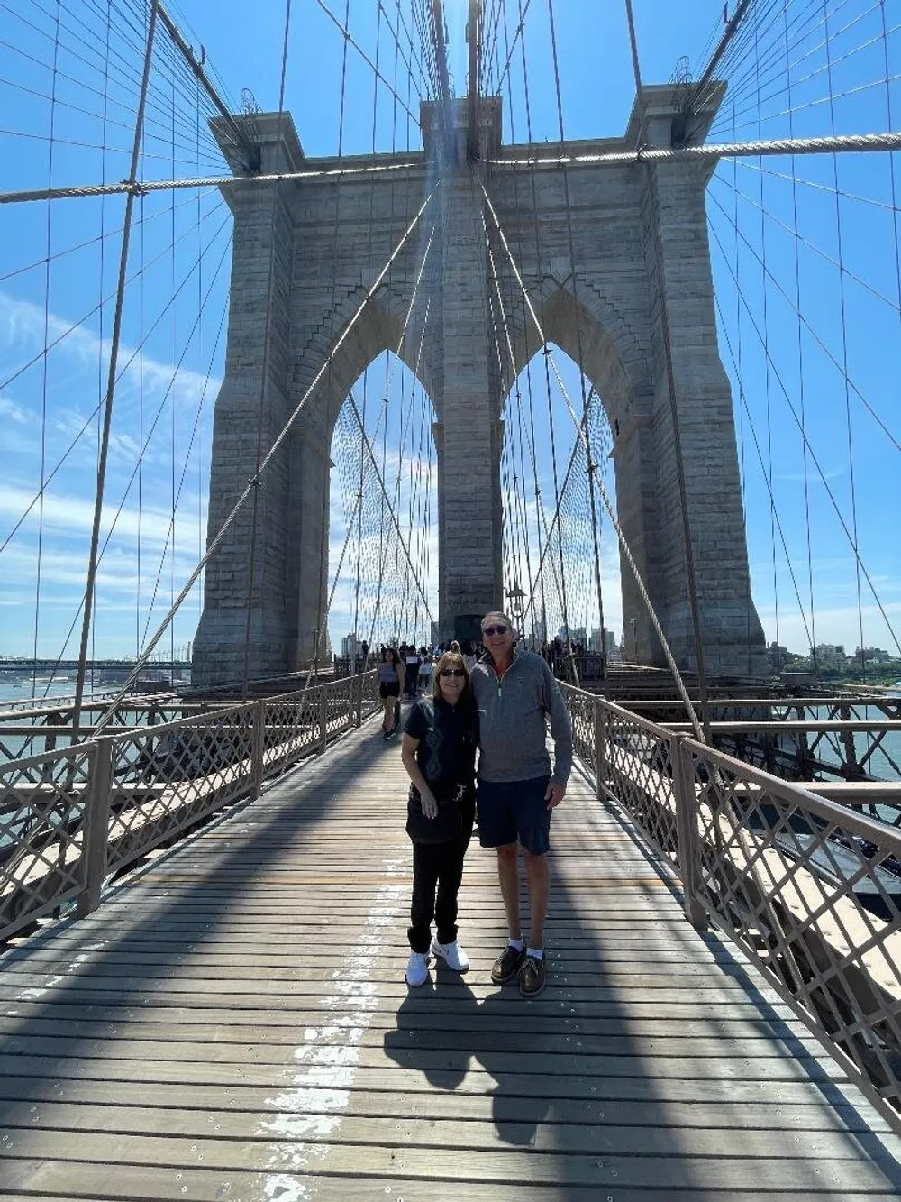 Two people are smiling for a photo on the walkway of a grand suspension bridge with intricate cables and stone towers under a clear blue sky