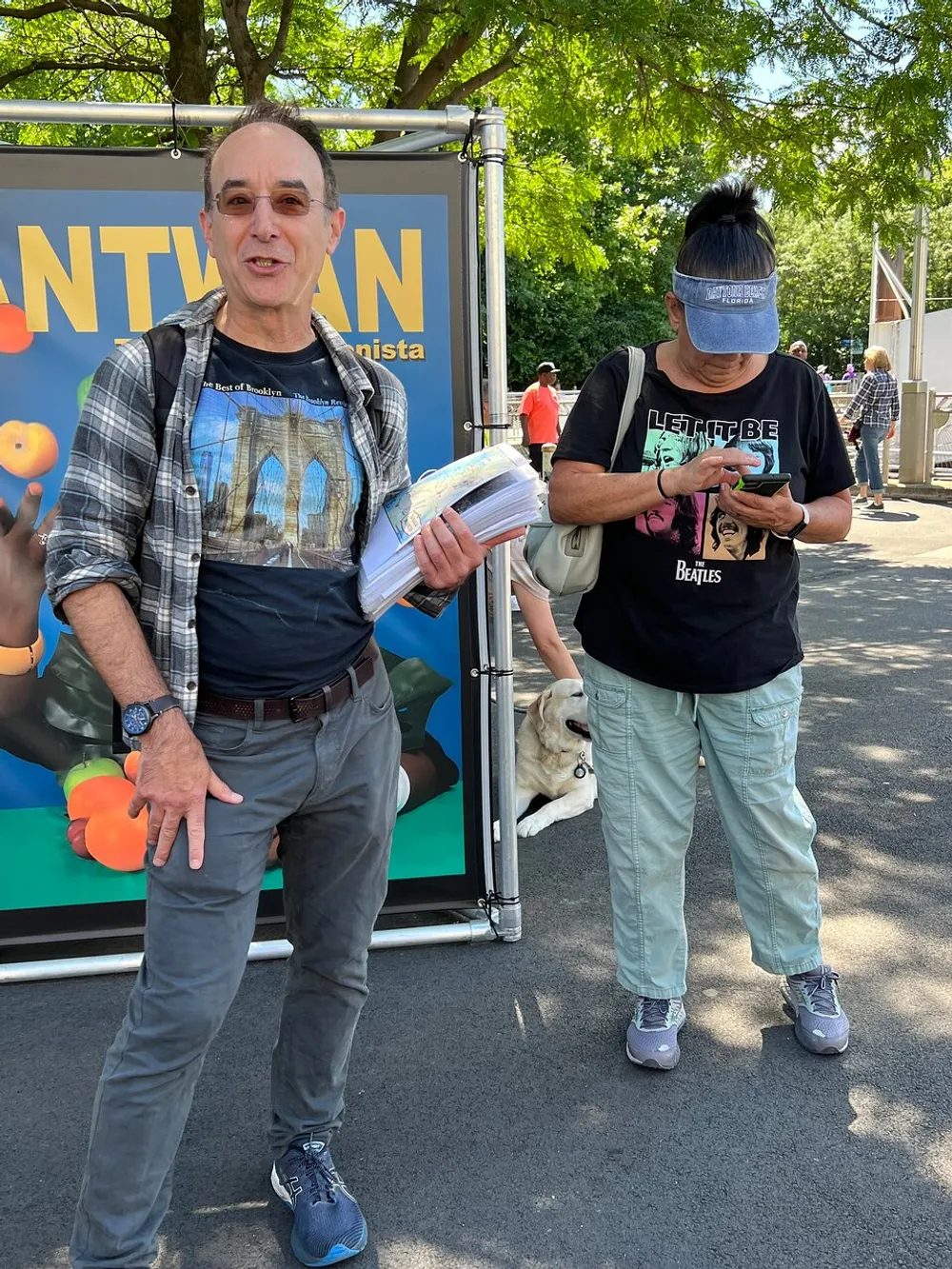A man and a woman are standing next to each other with the man holding a stack of papers and the woman looking at her phone there is a dog and a sign with the text ANTWAN visible in the background