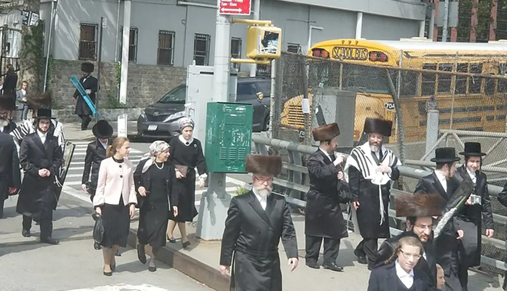 A group of individuals likely from a Jewish Orthodox community are walking down a street with a yellow school bus in the background