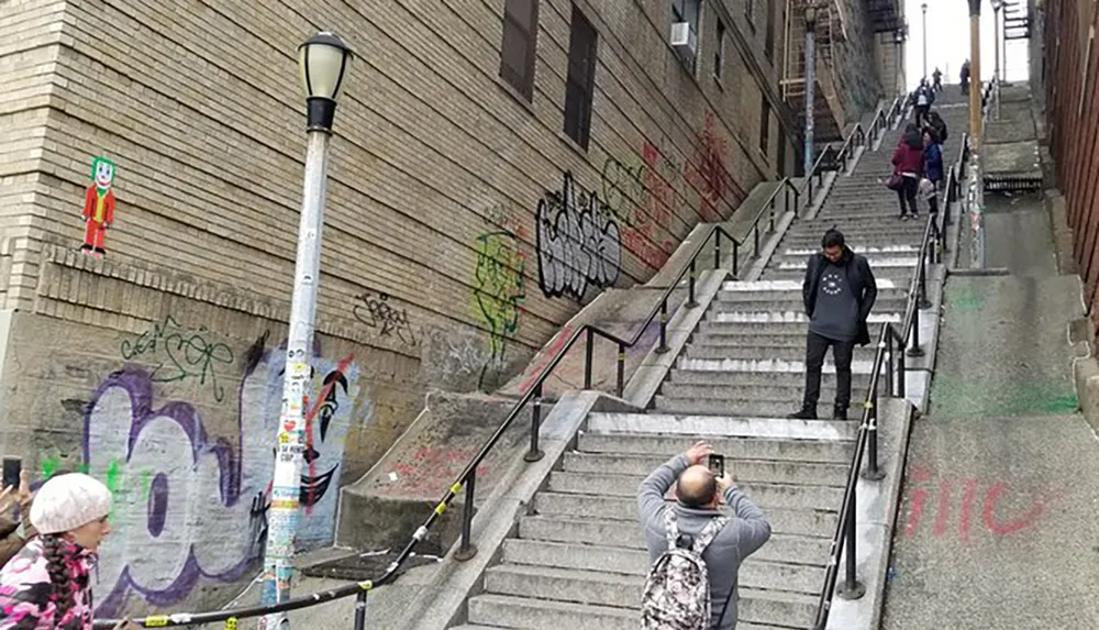 The image shows people on a graffiti-covered urban stairway with one individual taking a photo of another who is standing halfway up the stairs