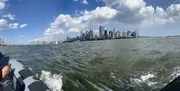 The image is a panoramic view of the Manhattan skyline as seen from a boat on the water, featuring clear skies and a few clouds above.