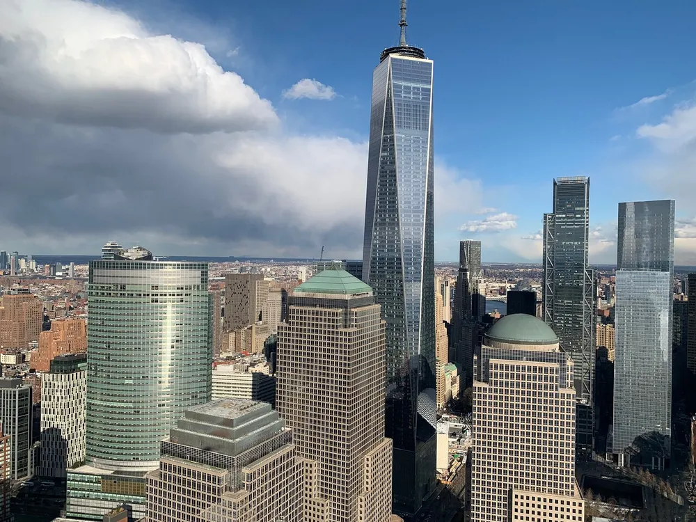 The image features a cityscape with several skyscrapers under a partly cloudy sky highlighting the One World Trade Center amidst the urban architecture