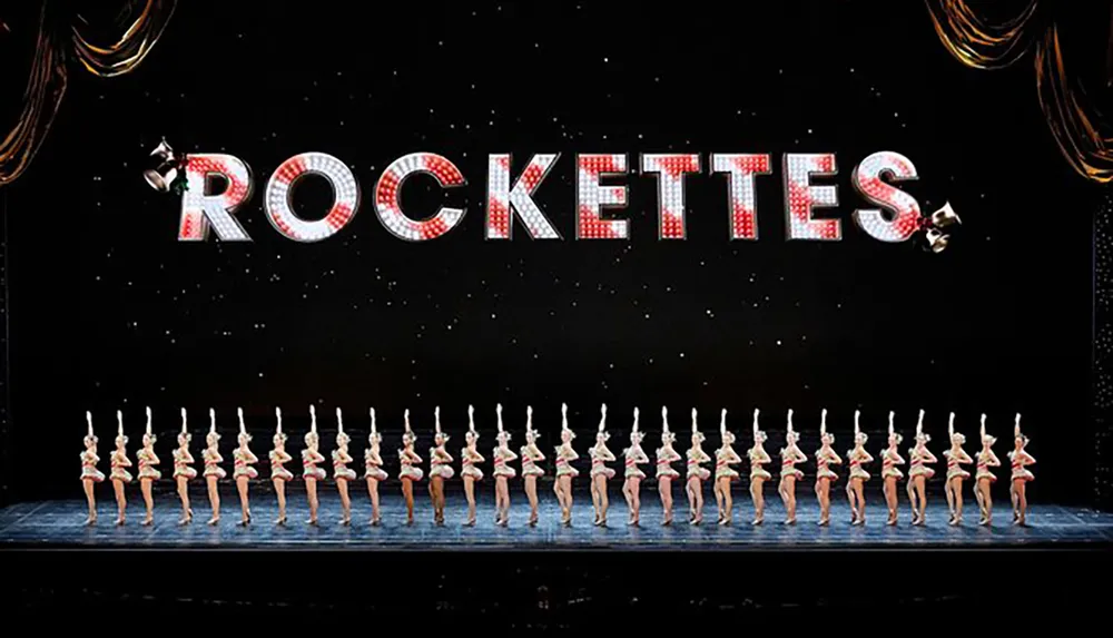 The image shows a line of performers in a coordinated dance pose on a stage beneath a large sparkling sign that spells out ROCKETTES