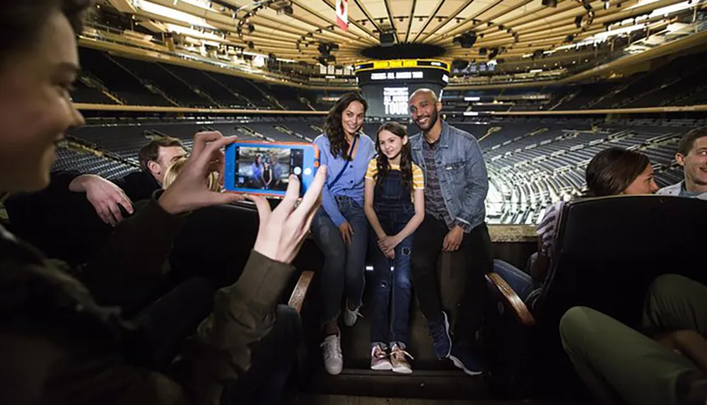 A person is taking a photo of two adults and a child smiling at the camera while sitting in an empty sports arena