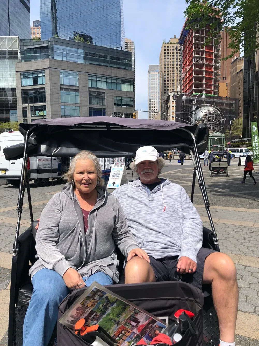 A smiling couple is sitting in a pedicab in an urban setting featuring modern buildings and a globe sculpture in the background
