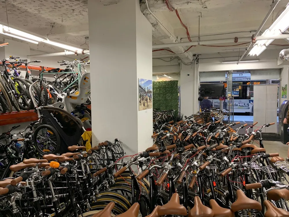 A crowded indoor bicycle parking area packed with numerous bikes with the view partially obstructed by a pillar and a glimpse of a city bus through the exit