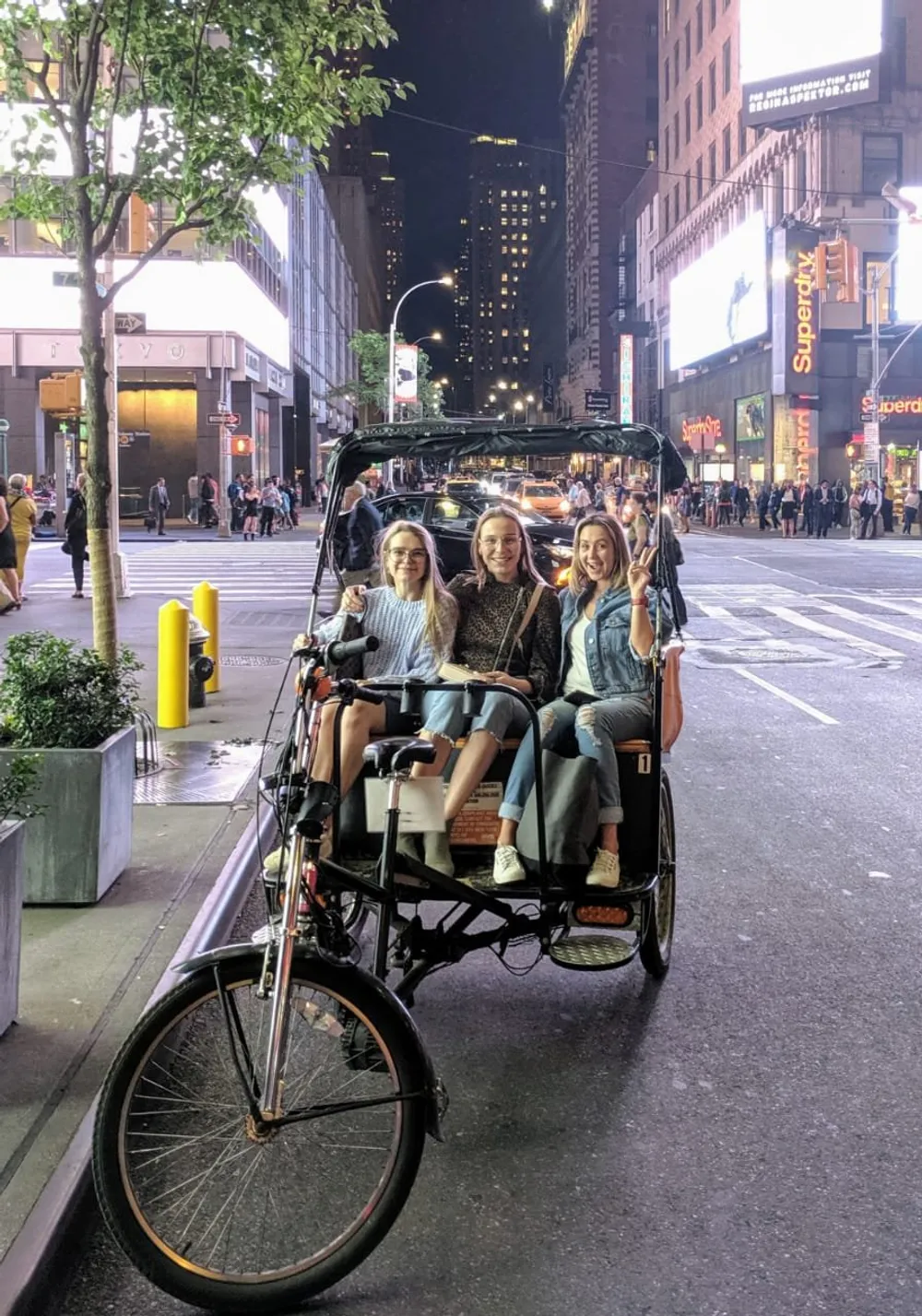 Three people are smiling while seated in a rickshaw on a brightly lit city street at night
