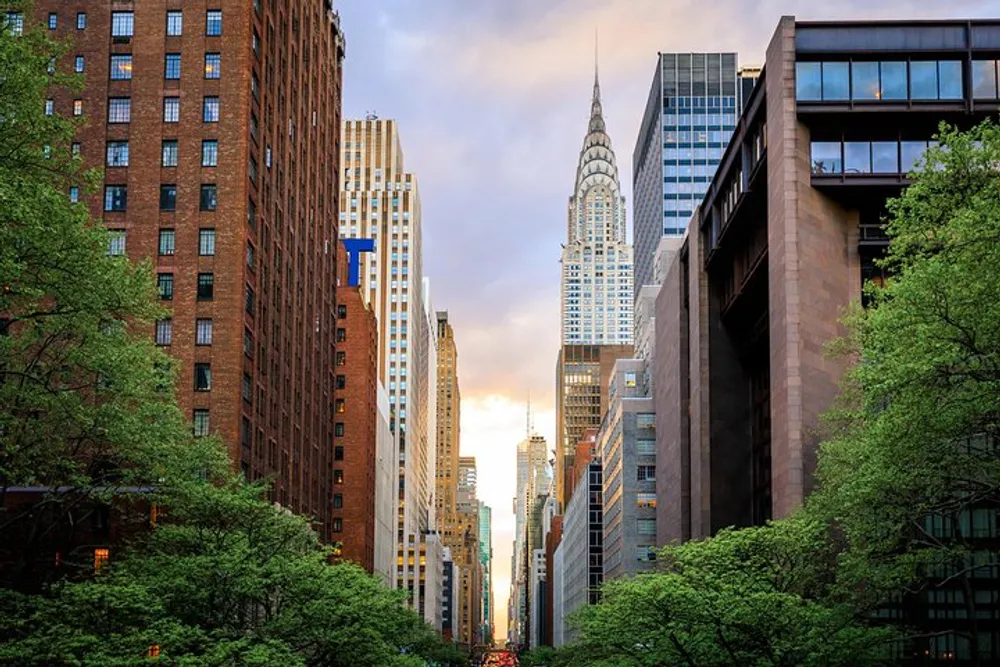 The image shows a view down a bustling urban street canyon lined with tall buildings leading towards an iconic skyscraper under a sky that is partly cloudy with a warm glow from the setting or rising sun