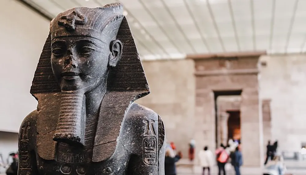 The image features an ancient Egyptian statue of a pharaoh exhibited in a museum with visitors in the background