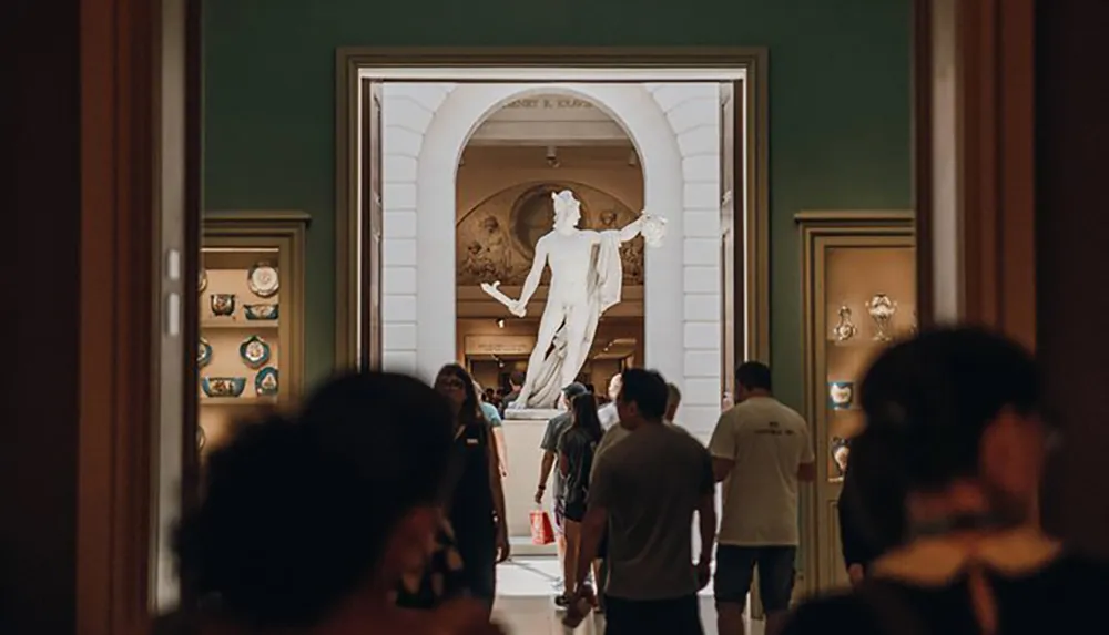 Visitors in a museum walk through galleries with walls adorned with art leading towards a central statue framed by archways