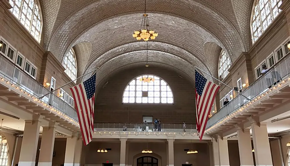 The image shows a spacious hall with a high arched ceiling adorned with two large American flags hanging from a balcony and a row of windows at the far end allowing natural light to flood in