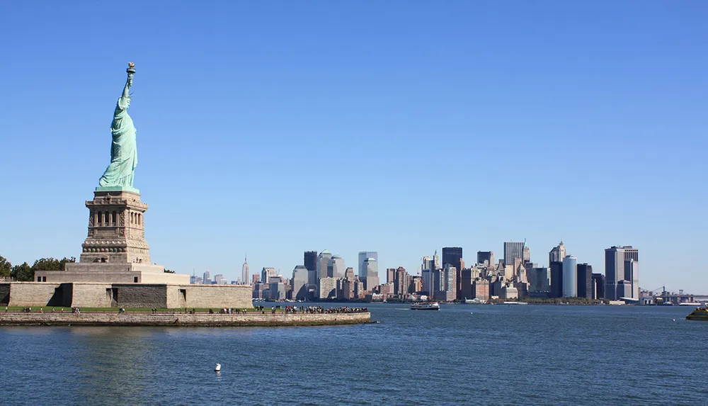 The image shows the Statue of Liberty in the foreground with the skyline of Lower Manhattan in the background under a clear blue sky