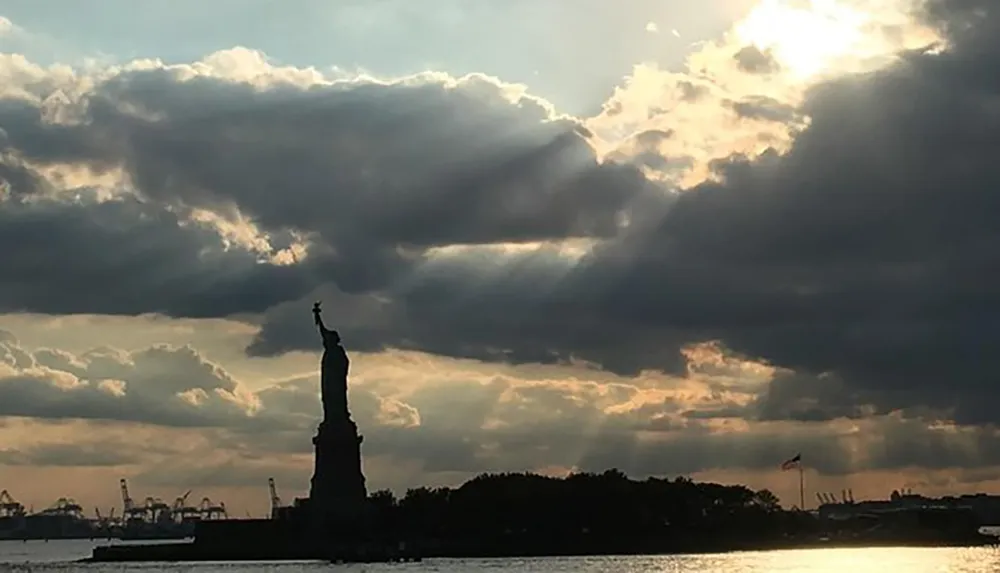The Statue of Liberty is silhouetted against a dramatic sky with sunbeams piercing through the clouds at dusk