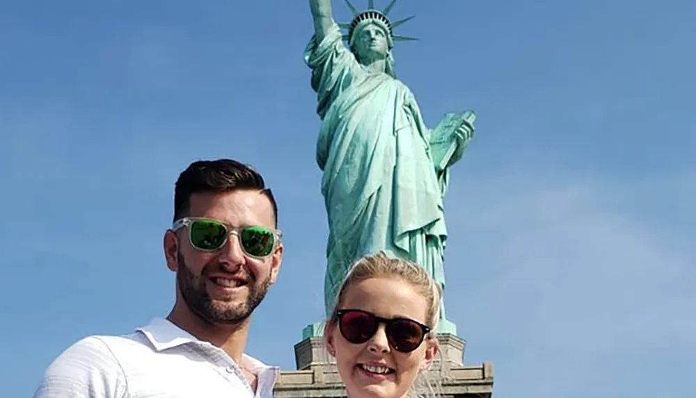 Two smiling individuals pose for a photo with the Statue of Liberty in the background under a clear blue sky