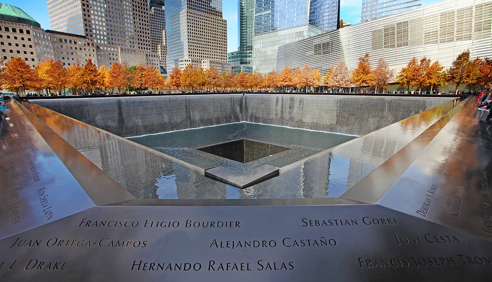 The image shows the reflecting pools at the National September 11 Memorial in New York surrounded by autumn-colored trees and skyscrapers with names inscribed around the edge in memory of the victims