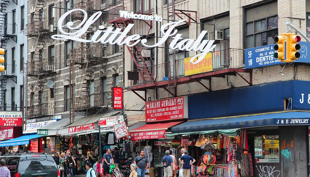 The image shows a bustling street scene in Little Italy with pedestrians shops and an iconic Welcome to Little Italy sign suspended above the sidewalk