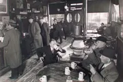 The black-and-white image captures a moment inside a bustling vintage tavern or cafe, where patrons clad in mid-20th-century attire engage in conversation, drink, and warm themselves by a large stove.