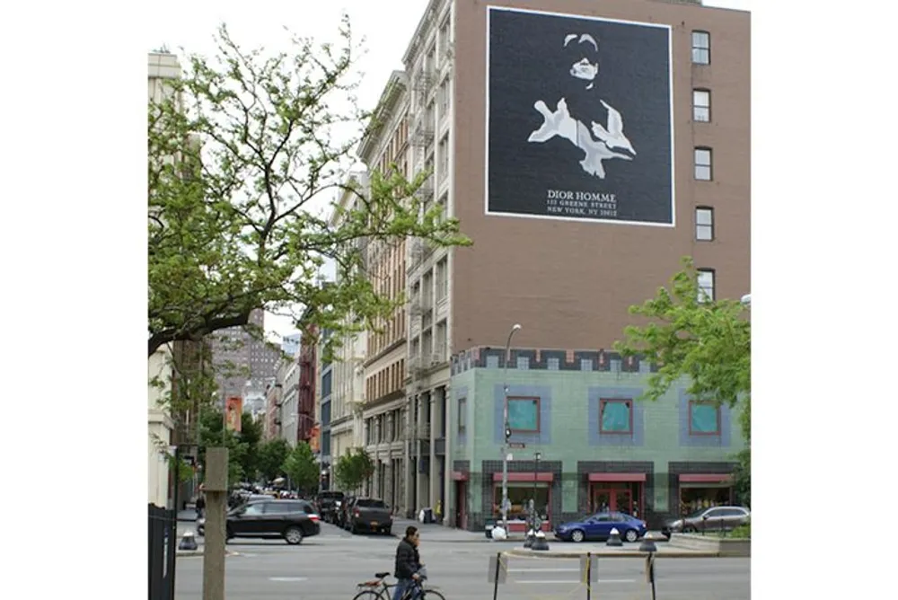 A person riding a bicycle on an urban street with a large Dior Homme advertisement featuring a shark on the side of a building