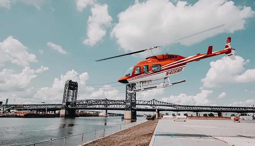 A red helicopter is flying near a bridge over a river under a partly cloudy sky