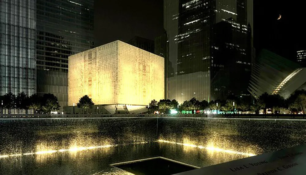 The image shows a luminous cube-shaped building at night near reflective pools with skyscrapers in the background and the partial view of an inscribed memorial edge in the foreground