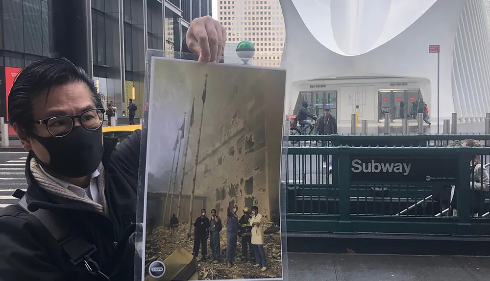 A person is holding up a photograph of responders at Ground Zero with the modern scene of the World Trade Center area in the background