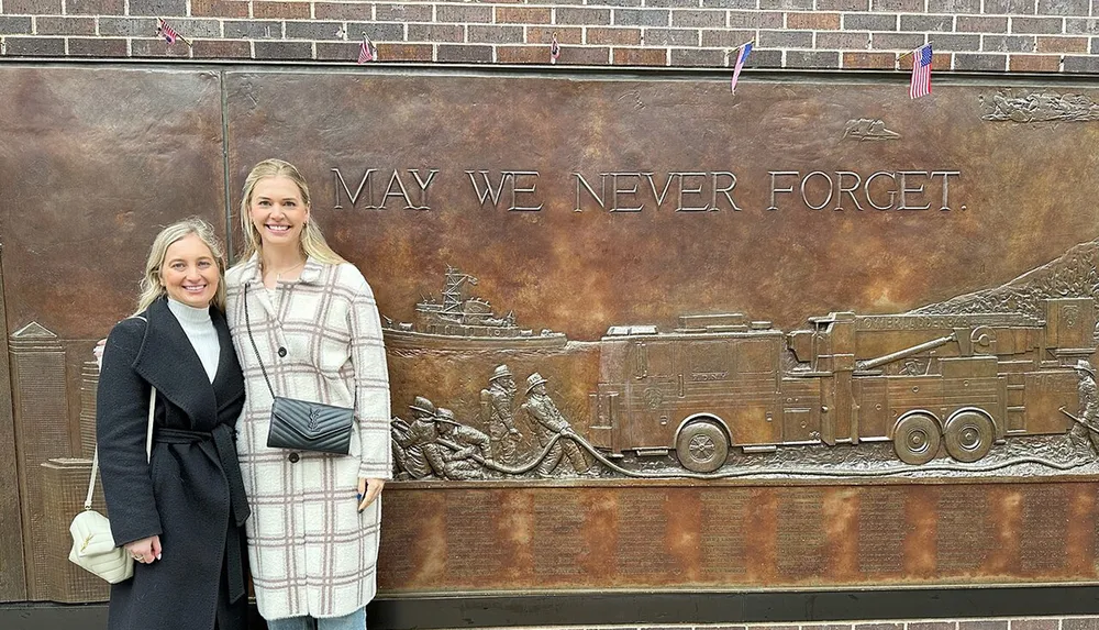 Two smiling women are standing in front of a memorial wall with a raised relief depicting firefighters in action and the phrase MAY WE NEVER FORGET inscribed on it