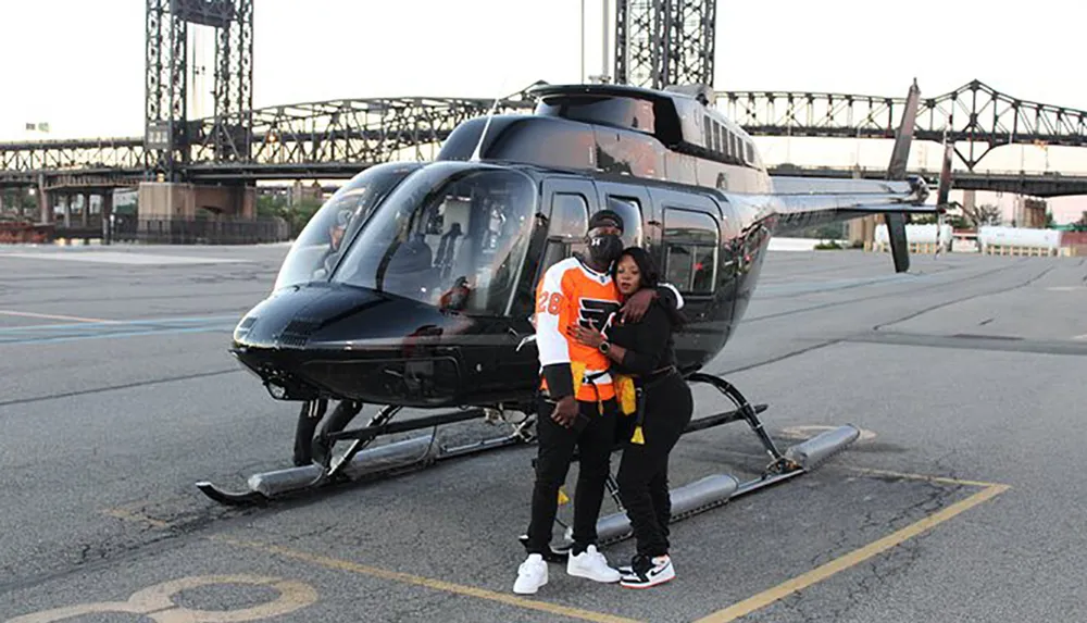 Two people are posing for a photo in front of a black helicopter on a landing pad with a bridge visible in the background