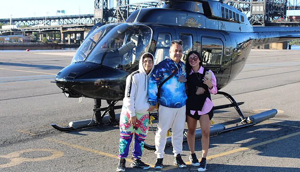 Three people are standing and smiling in front of a helicopter on a paved area with a bridge in the background