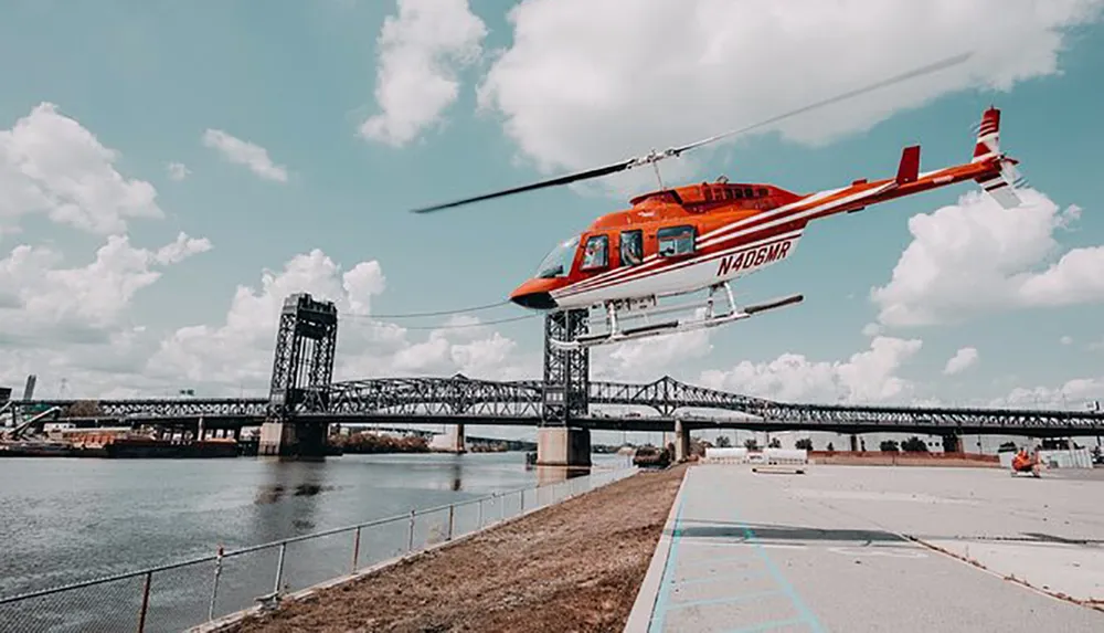 A red helicopter is in mid-flight near a bridge over a river with clouds in the sky