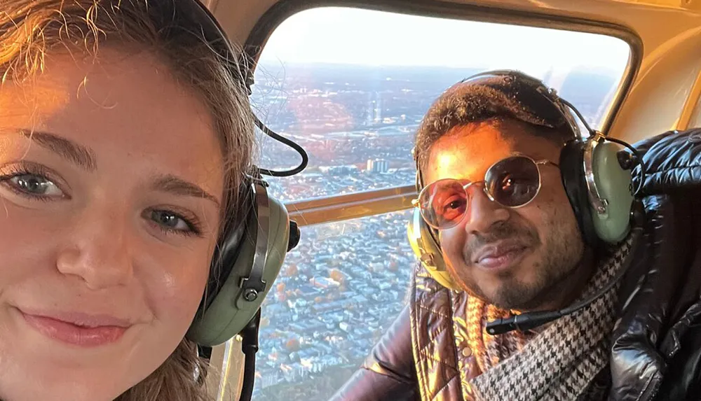 Two people are wearing headphones and smiling for a selfie inside a helicopter with a cityscape visible through the window in the golden light of sunset