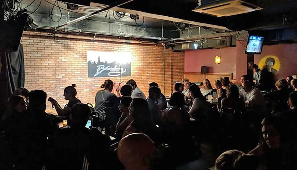 The image shows a dimly lit comedy club with a seated audience a stage in the background and a neon sign bearing the venues name on the brick wall