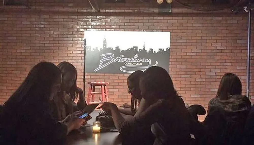 People are sitting in a dimly lit comedy club with a screen displaying The Broadway Comedy Club