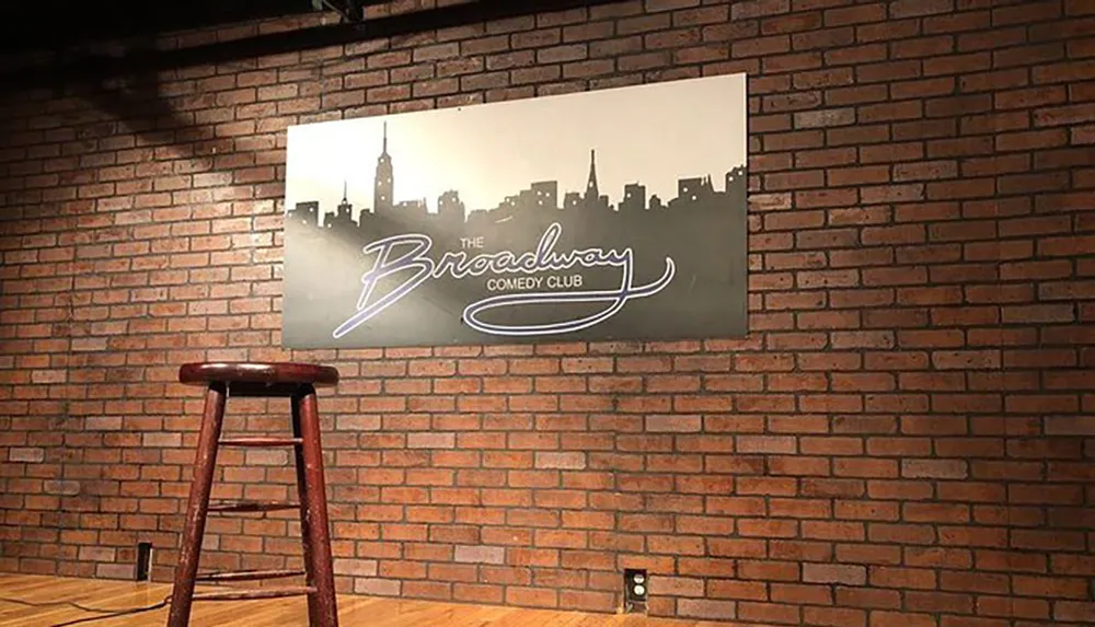 A single stool stands in front of a brick wall that features a sign for The Broadway Comedy Club suggesting a simple stage setup for stand-up comedy performances