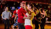 A group of people are joyfully engaging in social dancing at a vibrant venue with a couple in the foreground performing a dance move with eye contact and smiles.