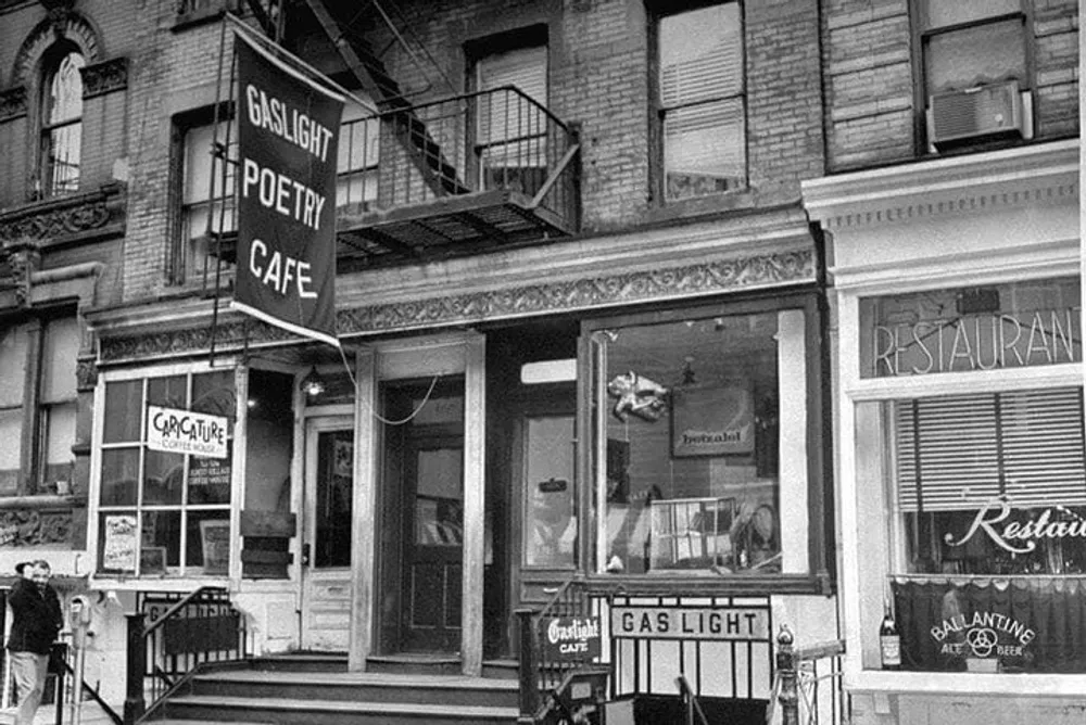 The image is a black and white photo of the exterior of the Gaslight Poetry Cafe complete with signs and a person standing by the entrance
